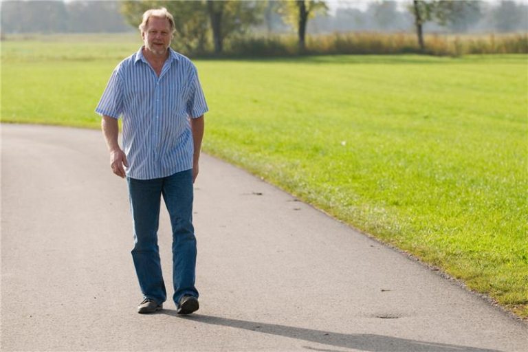 A person wearing a light blue striped shirt and blue jeans walking on a paved path in a park with green grass and trees in the background on a sunny day.