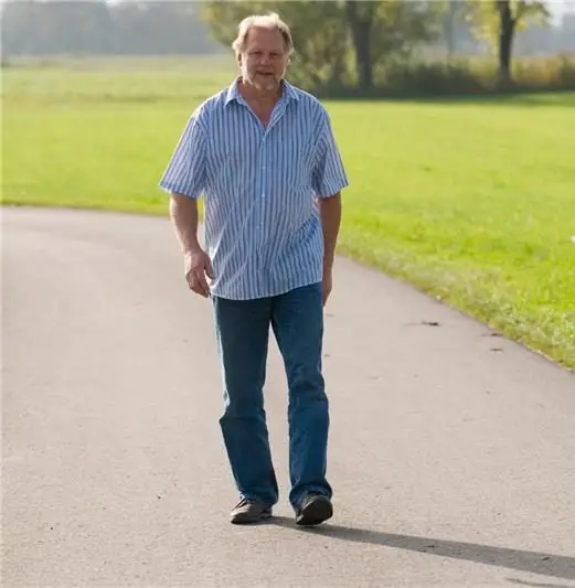 A person wearing a light blue striped shirt and blue jeans walking on a paved path in a park with green grass and trees in the background on a sunny day.