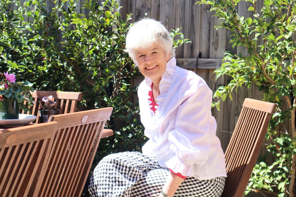 Wendy posing outside Wendy posing as she sits on an outdoor chair wearing a white blouse and smiling