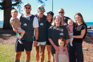 A group of seven people standing outdoors near the ocean, with tall trees and blue sky in the background. Two adults are holding young children, and several individuals are wearing matching dark gray shirts with the text “Run for Fun.”