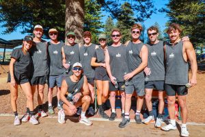 A group of eleven people wearing matching black athletic shirts with “Run for Mum” text, posing together outdoors on a sunny day. They are standing on a paved area surrounded by tall pine trees and holding water bottles.