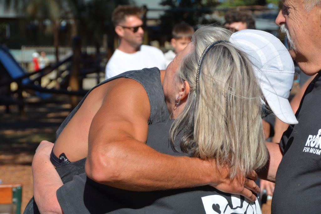 Will and his mum embracing outdoors after the charity run. Other participants and playground equipment are visible in the background.