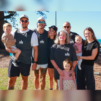 A group of seven people standing outdoors near the ocean, with tall trees and blue sky in the background. Two adults are holding young children, and several individuals are wearing matching dark gray shirts with the text “Run for Fun.”