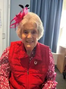 Rene Vivian smiles, wearing a pink floral shirt, red vest with a horseshoe pin, and a pink and black fascinator.