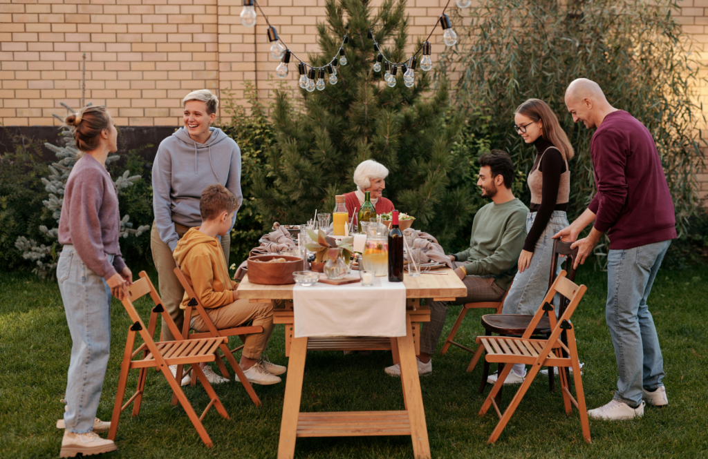 In a backyard setting, a family gathers around a wooden table set for a meal, with string lights overhead and a brick wall in the background.