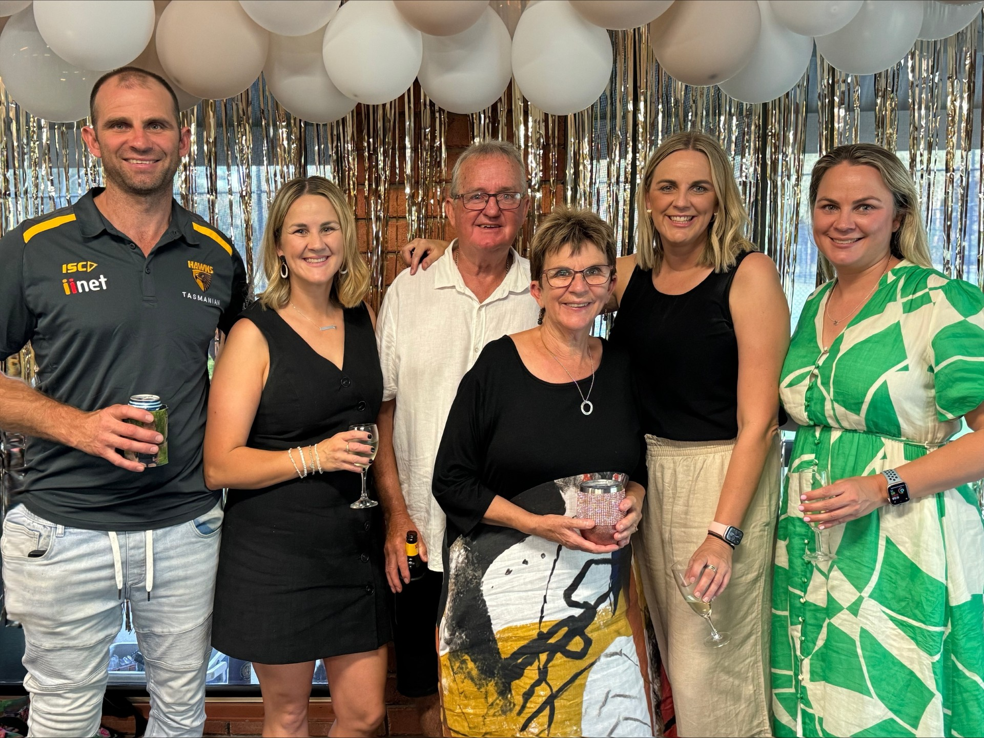 Megan's family including her dad gathered in front of a backdrop of white balloons and gold streamers, some holding drinks.