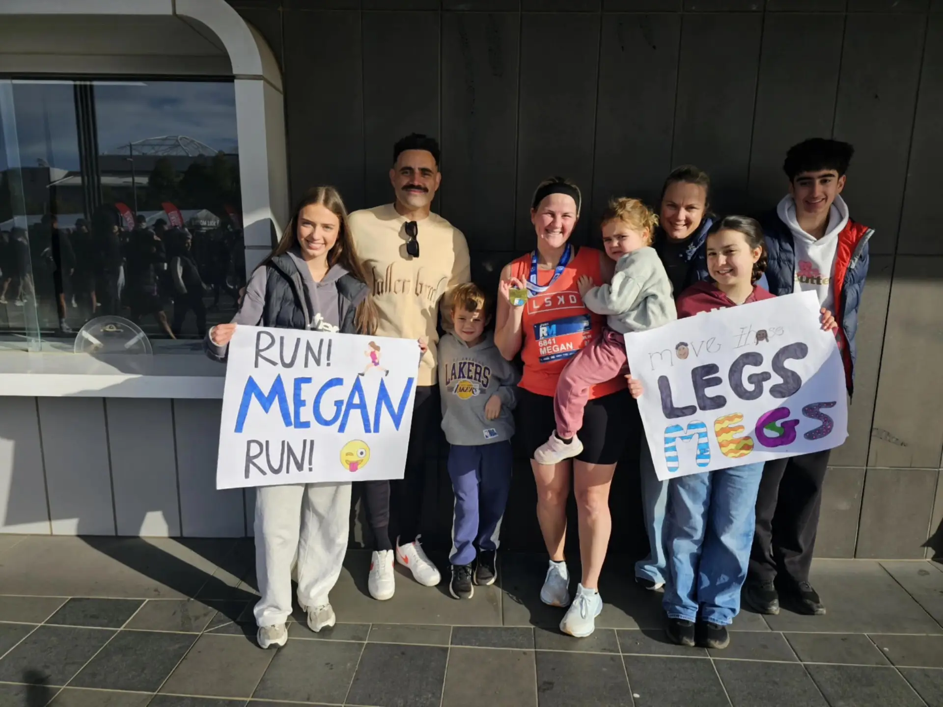 Megan's friends and family holding signs that say "Run Megan Run!" and "Move It Legs!" surround her in athletic wear and a medal, who is holding a young child.