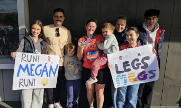Megan's friends and family holding signs that say "Run Megan Run!" and "Move It Legs!" surround her in athletic wear and a medal, who is holding a young child.