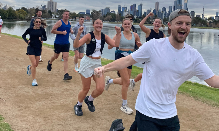 Colin running his final half marathon along a lakeside track, surrounded by supporters running beside him during the last push of his Seven in Seven challenge, with the city skyline in the background.