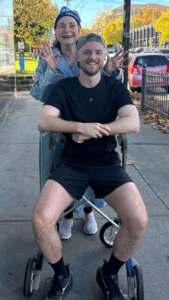 Colin sitting on a walker outdoors after one of his half‑marathons, while his friend and neighbour Isa stands behind him making a playful gesture, capturing a moment of support and camaraderie during his Seven in Seven challenge.