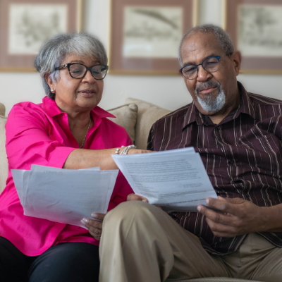 Two people sitting together on a couch reading printed documents, representing the process of reviewing and understanding advance care planning paperwork at home.