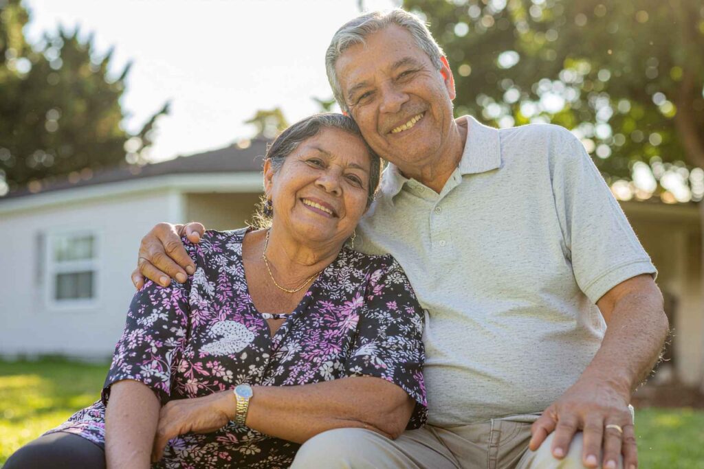 Elderly couple posing and smiling at the camera with a family house in the background