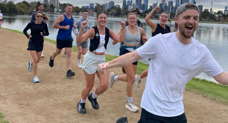 Colin running his final half marathon along a lakeside track, surrounded by supporters running beside him during the last push of his Seven in Seven challenge, with the city skyline in the background.