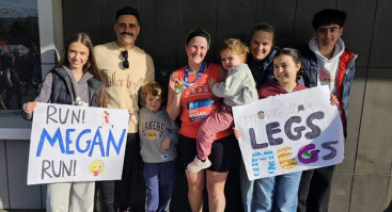 Megan's friends and family holding signs that say "Run Megan Run!" and "Move It Legs!" surround her in athletic wear and a medal, who is holding a young child.