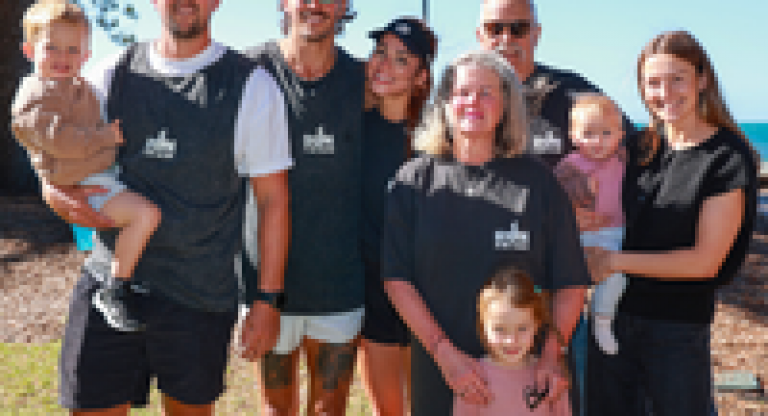 A group of seven people standing outdoors near the ocean, with tall trees and blue sky in the background. Two adults are holding young children, and several individuals are wearing matching dark gray shirts with the text “Run for Fun.”